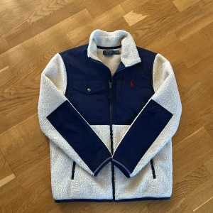 Vit och marinblå teddyjacka Polo Ralph Lauren - Snygg teddyjacka från Polo Ralph Lauren i vit och marinblå färg. Jackan har dragkedja, bröstficka med knapp och två sidofickor. Materialet är mjuk fleece med förstärkta partier i polyester. Perfekt för kalla dagar och riktigt stilren look. Ny pris 2800 mitt pris 1400