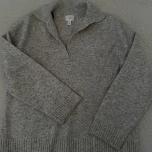 Grå stickad tröja från H&M - Mysig grå stickad tröja från H&M med krage och v-ringad öppning. Tröjan har långa ärmar och breda ribbade muddar vid ärmslut och nederkant. Perfekt för lager-på-lager och chill dagar.