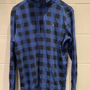 Blårutig half zip från Polo Ralph Lauren - Snygg långärmad tröja från Polo Ralph Lauren i blått och svart rutigt mönster. Tröjan har half zip framtill och klassisk broderad logga på bröstet. Tillverkad i mjuk bomull, perfekt för lager-på-lager under höst och vinter. Katt finns i hemmet.
