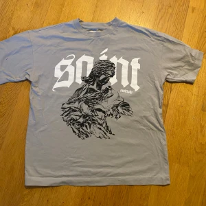 Grå t-shirt Multiply Apparel S - Grå t-shirt från Multiply Apparel i storlek S med stor vit och svart print framtill. Trycket har ett konstnärligt motiv och texten 'saint' i gotisk stil. T-shirten har rund hals och korta ärmar, tillverkad i mjuk bomull. Perfekt för dig som gillar streetwear och grafiska prints. Den har en liten fläck men syns knappt.