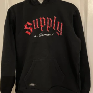 Svart hoodie Supply & Demand X-Large - Svart hoodie från Supply & Demand med röd broderad text och vit text på bröstet. Hoodien har mönstrat innerfoder i luvan, känguruficka och snörning vid huvan. Perfekt för dig som gillar streetwear vibes. Storlek XL för barn. Passar oxå M. Ny. 