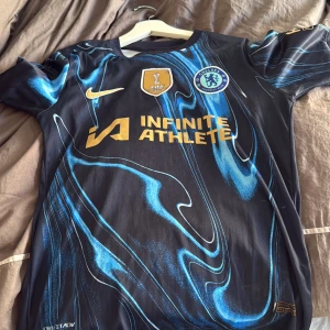 Chelsea FC Palmer 20 bortatröja Nike - Chelsea FC bortatröja med Palmer 20-tryck, tillverkad av Nike. Tröjan är svart med blått abstrakt mönster och har klubbmärke, FIFA-märke och sponsorlogga i guld. Kortärmad modell i lätt funktionsmaterial, perfekt för match eller träning.