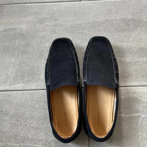 Mörkblå mockaloafers från Gant - Snygga mörkblå loafers från Gant i mocka med kontrasterande vita sömmar. Skorna har en klassisk, stilren design med fyrkantig tå och platt sula. Perfekta för dig som gillar en clean och tidlös look.