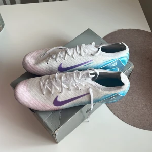 Nike fotbollsskor vit/blå/lila - Säljer ett par Nike fotbollsskor med snyggt gradientmönster i vitt, blått och lila. Skorna har lila swoosh, snörning och en cool metallicblå sula med dobbar. Ovandelen är i lätt syntetmaterial för maxad känsla på planen. Skröna sitter lite som 44/45 