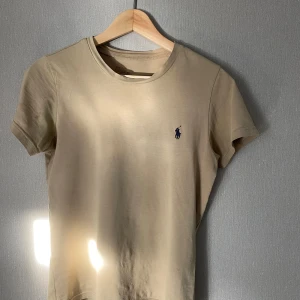 Beige t-shirt från Polo Ralph Lauren - Snygg beige t-shirt från Polo Ralph Lauren med klassisk rund hals och korta ärmar. Den är i storlek S men är ganska tajt i passformen så man kan säga att den är i storlek XS. Den har en liten fläck från något jag inte vet vad det är, men som tur är den endast synlig när man kollar på den på nära håll. Annars har den helt perfekt skick👌