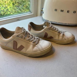 Veja vita sneakers med rosa detaljer - Snygga Veja sneakers i vitt skinn med ljusrosa V-logga och hälparti. Klassisk låg modell med rund tå, platt sula och snörning. Perfekta för dig som gillar stilrena och trendiga skor med en twist. Passar till allt och ger en clean look.