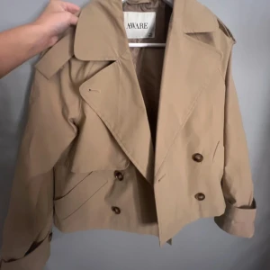Beige kort trenchcoat från AWARE - Snygg beige trenchcoat från AWARE med dubbelknäppning, bred krage och axelklaffar. Jackan och stora knappar samt klassiska detaljer som gör den extra trendig. Perfekt för dig som vill ha en stilren och modern look. XS men passar mig som bär S. säljer då den inte kommer till användning ❤️❤️