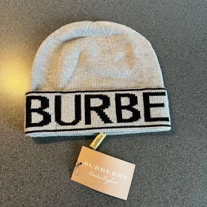 Burberry mössa med logga - Snygg mössa från Burberry med bred uppvikt kant och stor svart logotyp. Finns i både ljusgrått och beige, båda med svart text. Klassisk och stilren design i stickat material, perfekt för att hålla värmen och ge en exklusiv touch till din outfit.