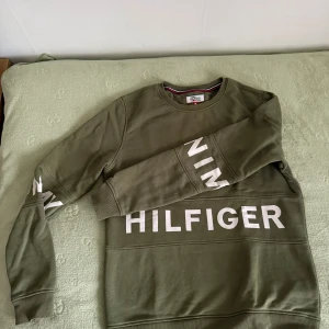 Olivgrön sweatshirt Tommy Hilfiger - Olivgrön sweatshirt från Tommy Hilfiger Denim med stora vita tryckta orden ”tommy” och ”denim” runt ärmarna och ”hilfiger” runt brösten. Tröjan har rund halsringning, ribbade muddar och är tillverkad i mjuk bomull. Perfekt för en avslappnad och stilren look.