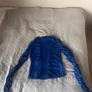 Blå långärmad topp från Gina Tricot - Säljer en blå långärmad topp från Gina Tricot i storlek S. Toppen har snygga rynkade detaljer längs ärmarna och framtill, vilket ger en cool och trendig look. Tillverkad i ett stretchigt och glansigt polyestermaterial som sitter skönt på kroppen.