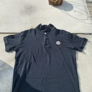 Mörkblå Moncler piké t-shirt - Svart piké t-shirt från Moncler med klassisk krage och knappslå framtill. T-shirten har korta ärmar och Moncler-logga på bröstet. Tillverkad i mjuk bomull för en skön känsla. Perfekt för dig som gillar stilrena och exklusiva plagg.