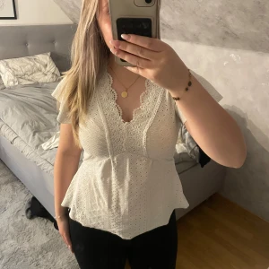 Vit spetsblus med volangärm - Säljer en superfin vit blus med v-ringning och spetsdetaljer framtill. Blusen har korta volangärmar och är tillverkad i bomull med broderade hålmönster. Den har en lätt utsvängd passform nedtill som ger en feminin siluett. Perfekt för dig som gillar romantisk och söt stil.