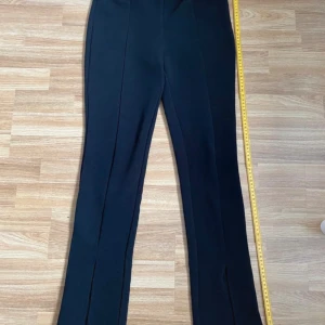 Kostymbyxa - byxa med slits - Eleganta svarta kostymbyxor med normalhög (29 cm) midja och en smickrande tajt passform. Resår i midjan som mäter 4,5 cm.  Byxorna har en subtil slitsdetalj (18,5 cm) nedtill i båda benen som ger en modern touch. Perfekta för både vardag och fest. Tvättrådslappen är bortklippt. Därav är min troliga gissning att materialet är polyester.  Annars inga defekter.
