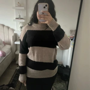 Randig stickad tröja i beige och svart - Mysig stickad tröja med breda svarta och beige ränder. Tröjan har en relaxed fit, rund halsringning och långa ärmar. Perfekt för att hålla sig varm och snygg under kyliga dagar.