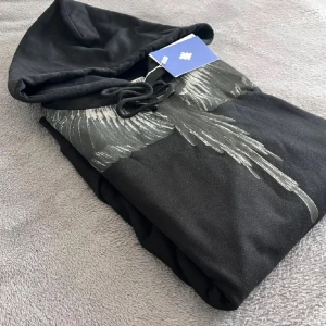 Svart hoodie Marcelo Burlon med vingar - Svart hoodie från Marcelo Burlon County of Milan med ikoniskt vingtryck i grått över axlar och bröst. Tröjan har huva med snörning, känguruficka och är tillverkad i mjuk bomull. Perfekt för dig som vill sticka ut med en streetstyle vibe.