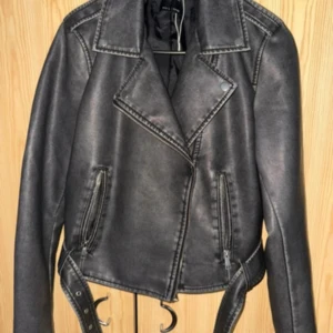 Svart skinnjacka från Sinsay, stl L - Cool svart skinnjacka från Sinsay i storlek L. Jackan har klassisk bikerstil med bred krage, dragkedjor på fickorna och bälte i midjan. Snygga detaljer och en lite croppad passform som ger en edgy vibe till din outfit.