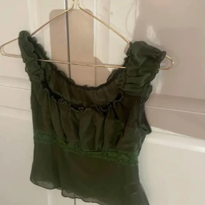 Olivgrön blus med volangdetaljer - Säljer en olivgrön blus i transparent material med breda volangprydda axelband och rynkade detaljer längs halsringningen. Blusen har en spetskant under bysten som ger en extra touch. Perfekt för dig som gillar romantisk och trendig stil.