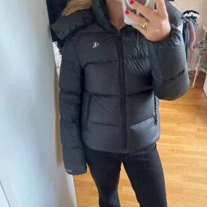 Svart dunjacka Calvin Klein Jeans XS - Snygg svart dunjacka från Calvin Klein Jeans i storlek XS. Jackan har dragkedja framtill, två fickor med dragkedja och hög krage. Tillverkad av återvunnen nylon och polyester med ansvarsfullt dun. Perfekt för kalla dagar och riktigt stilren look.