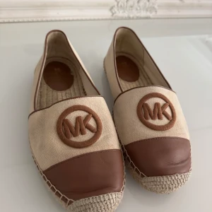 Beige och bruna Michael Kors loafers - Snygga loafers från Michael Kors i beige canvas med bruna skinn-detaljer och stort MK-logo framtill. Sulan är flätad och ger en avslappnad vibe. Perfekta för dig som gillar stilrena och trendiga skor med en touch av lyx.