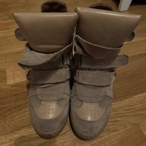 Isabel marant liknande - Snygga beige sneakers från Carinii med kilklack och breda kardborreband. Skorna är gjorda i mocka och skinn, har rund tå och coola detaljer på tungan. Perfekta för dig som vill ha lite extra höjd. Folk har trott att dem är riktiga på mig då de är så lika orginalet. pris kan absolut diskuteras!🫰