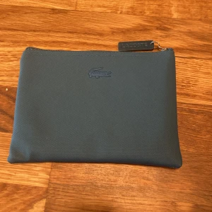 Blå kuvertväska från Lacoste - Snygg blå kuvertväska från Lacoste med klassisk krokodil-logga framtill. Väskan är tillverkad i tålig syntet och har en stilren design med dragkedja upptill. Perfekt för att bära det viktigaste och ger en clean vibe till din outfit.