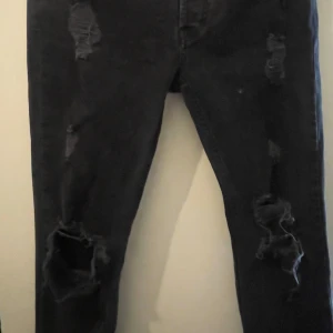 Svarta ripped jeans från Tigha - Säljer ett par svarta jeans från Tigha i modellen Morten. Jeansen har flera slitningar och stora hål på knäna för en riktigt edgy look. De är i stretchigt denim och har klassisk femficksdesign. Perfekta för dig som gillar streetstyle.