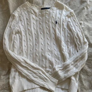 Vit kabelstickad tröja Polo Ralph Lauren - Kabelstickad vit tröja från Polo Ralph Lauren med klassisk rund halsringning och broderad logga på bröstet. Tröjan är i bomull och har långa ärmar samt ribbade muddar för en snygg och tidlös look.