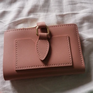 Rosa plånbok med gulddetaljer - Söt rosa plånbok i syntetmaterial med guldfärgade detaljer och dragkedja. Plånboken har flera kortfack, ett fönster för ID och en praktisk ficka med tryckknapp på framsidan. Perfekt storlek för att få plats med allt viktigt i väskan.                                      Plånboken är från Glitter och är endast använd en gång. 