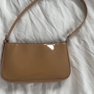 Beige handväska i glansigt material - Snygg beige/brun handväska i glansigt syntetmaterial med enkel, stilren design. Väskan har en smal axelrem och stängs med dragkedja upptill. Perfekt storlek för det viktigaste och passar till många olika outfits.