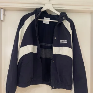 Blå och vit vindjacka från Bershka - Säljer en snygg vindjacka från Bershka i blå och vitt med coola kontrastpaneler och trycket 'Controlled Chaos' på bröstet. Jackan har huva, meshfoder och dragkedja framtill. Perfekt för dig som gillar streetstyle och sportig vibe.