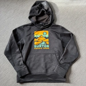 Burton - Svart Burton hoodie med tryck - Svart hoodie från Burton med färgglatt tryck framtill som visar berg, sol och vatten. Tröjan har huva, magficka och tumhål i ärmsluten. Perfekt för dig som gillar streetwear och outdoor vibes. Ett litet hål (slitage) finns längst nere på slutet av högra ärmen, i övrigt inga defekter. 