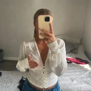 Vit blus med knappar och blommönster - Säljer en vit blus med djup v-ringning, långa puffärmar och dekorativa knappar framtill. Blusen har ett subtilt blommönster i tyget och är figurnära i midjan. Perfekt för dig som gillar romantiska detaljer och en feminin look. Från VENDERBYS! 