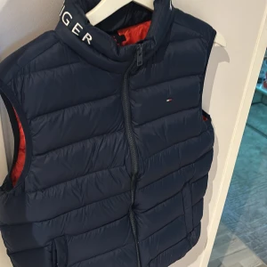 Blå dunväst från Tommy Hilfiger - Snygg blå dunväst från Tommy Hilfiger med röd insida och dragkedja framtill. Västen har en hög krage med märkets logga och är perfekt för kyligare dagar. Den quiltade designen ger en stilren look.