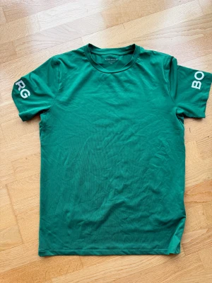 Grön Björn Borg t-shirt - Snygg grön t-shirt från Björn Borg med tryckt logga på båda ärmarna. Klassisk rund hals och korta ärmar. Perfekt för dig som gillar sportiga och stilrena plagg.