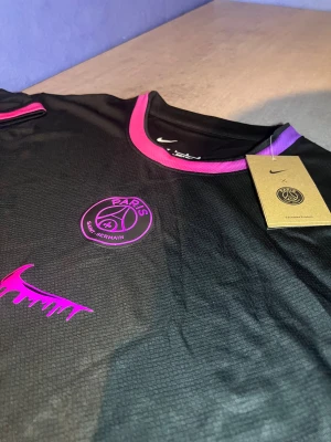 PSG x Nike svart t-shirt med lila detaljer - Svart PSG x Nike t-shirt med lila och rosa detaljer på krage och ärmslut. Snyggt Paris Saint-Germain-emblem och Nike-logga i lila på bröstet. Tillverkad i lätt polyester som andas, perfekt för fotboll eller chill. Modern och sportig look.
