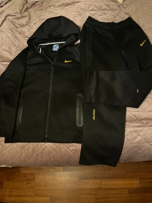 Nike NOCTA dress/set - Svarta mjukisbyxor från Nike NOCTA med gul logga på benet och gul Swoosh vid höften. Byxorna har dragsko i midjan, zip-försedda fickor och en loose passform. Materialet är mjukt och skönt, perfekt för chill eller träning. Snygga detaljer med NOCTA-tryck på benet.