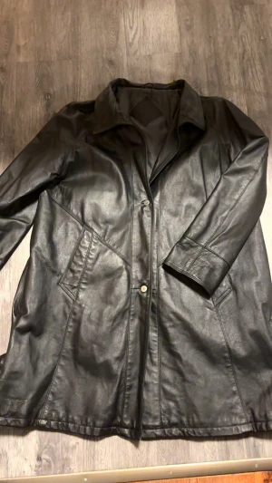 Svart skinnrock från Goldstein - Svart lång skinnrock från Goldstein by Molltan med klassisk krage och två snedställda fickor framtill. Jackan har knäppning med knappar och stilrena sömmar som ger en snygg siluett. Perfekt för dig som gillar tidlös och edgy stil.