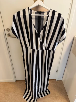 Svartvit jumpsuit från SHEIN - Jumbsuit från SHEIN med svartvita vertikala ränder, v-ringad omlott framtill och korta ärmar. Tillverkad i lätt polyester som ger ett snyggt fall. Dragkedja i ryggen för enkel påklädning. Perfekt för dig som gillar statement-plagg med grafisk känsla.