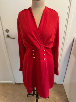 Röd omlottklänning från SHEIN XL - Röd kort omlottklänning från SHEIN med långa ärmar, markerad midja och dekorativa guldknappar framtill. Klänningen har v-ringning och stängs med dragkedja bak. Tillverkad i ett mjukt, lätt material som ger en snygg siluett.