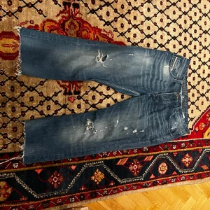 Blå jeans med slitningar - Säljer ett par blå jeans i storlek 38 med coola slitningar på framsidan. De har en rak passform och fransiga benslut för en avslappnad look. Perfekta för en casual stil och tillverkade i Italien. Passar bra till både vardag och fest!
