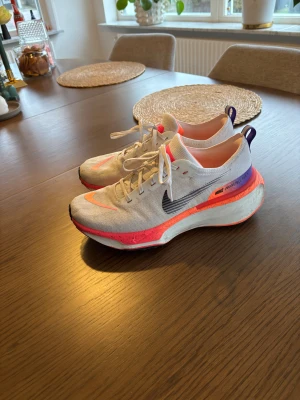 Nike ZoomX Invincible Run sneakers - Nike ZoomX Invincible Run sneakers i vitt med neonorange och lila detaljer. Skorna har en tjock, dämpad sula och svart swoosh på sidan. Ovandelen är i mesh-liknande syntetmaterial för bra andningsförmåga. Perfekta för löpning och träning, med snörning och cool design.