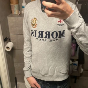 Morris xs  - Snygg grå sweatshirt från Morris Gents i storlek XS. Tröjan har broderade detaljer med brittisk flagga och emblem på bröstet samt texten 'Morris The Gent' i mörkblått. Klassisk rund halsringning och ribbade muddar passar 165-172 ungefär 