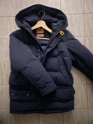 Mörkblå Parajumpers pufferjacka - Säljer en mörkblå pufferjacka RUGGED från Parajumpers med huva, flera fickor och snygga detaljer som gul snörning och patch på ärmen. Jackan har quiltad design och dragkedja framtill. Perfekt för kalla dagar och har en cool, urban look.