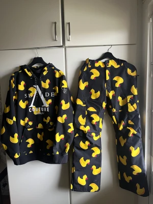 Slade täckbyxor med gummiankor - Unika täckbyxor från Slade med heltryck av gula gummiankor på svart bakgrund. Byxorna har cargofickor och bred passform, perfekta för dig som vill sticka ut i backen. Materialet är tåligt och vattenavvisande polyester. Snygg och lekfull design som verkligen syns.