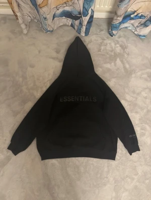 Svart Essentials Fear of God hoodie - Säljer en svart hoodie från Essentials Fear of God med stor huva och tryck på bröstet samt ärmen. Hoodien har en klassisk känguruficka framtill och ribbade muddar vid ärmslut och nederkant. Materialet är mjukt och tjockt, perfekt för chill dagar.