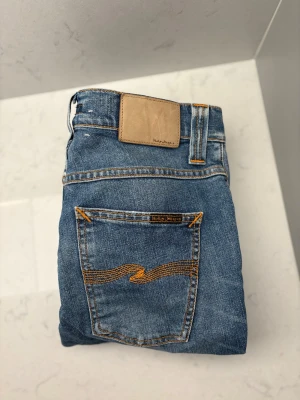 Blå jeans från Nudie Jeans - Snygga blå jeans från Nudie Jeans med klassisk orange söm på bakfickorna och en ljusbrun patch i midjan. Jeansen har en rak passform och är tillverkade i slitstarkt denimtyg. Perfekt för dig som gillar tidlös stil och coola detaljer.