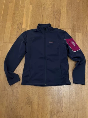 Blå vindjacka Peak Performance XL - Snygg marinblå vindjacka från Peak Performance med rosa detalj och ficka med dragkedja på ärmen. Jackan har hög krage, hel dragkedja framtill och sportig design. Perfekt för dig som gillar stilrena och funktionella plagg.