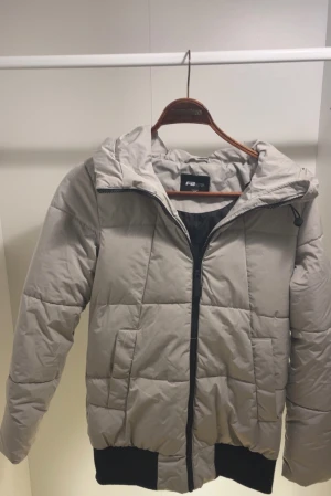 Beige puffjacka från FB Sister – storlek XS  - Snygg och varm puffjacka i beige med svart innerfoder och svarta detaljer. Har huva, dragkedja framtill och två sidofickor. Resår längst ner för bättre passform. Använd få gånger och i mycket fint skick, inga fläckar eller skador.