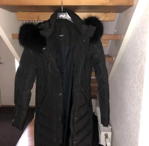 Svart lång dunjacka med fuskpäls - Lång svart dunjacka med stor huva och fluffig svart fuskpäls runt kragen. Jackan har dragkedja och knappar framtill samt två fickor med dragkedja. Perfekt för kalla dagar och riktigt snygg siluett.