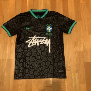 Brasil fotbollströja - Unik svart Brasil-fotbollströja från Stüssy. Tröjan har korta ärmar, gröna och blå detaljer på krage och ärmslut, samt ett subtilt leopardmönster. Stor Stüssy-logga framtill och Brasil-märke på bröstet. Perfekt för dig som gillar streetwear och fotboll.